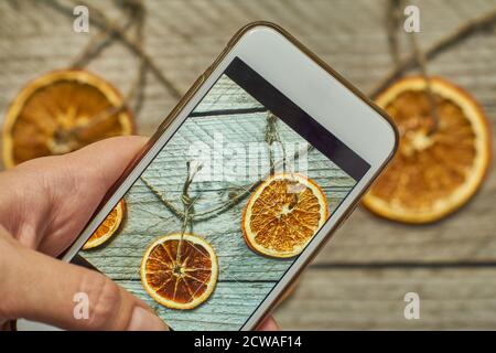 Frau macht ein Foto von weihnachtsdekoration aus getrockneten Orangenscheiben auf ihrem weißen modernen Smarthfone. Unter Blogging conent für weihnachten und neue y Stockfoto