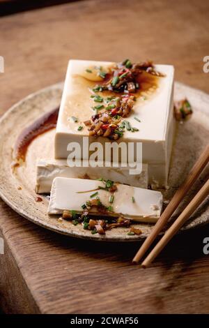 Seidentofu japanischer Sojakäse Vollstück mit Chili, Ingwer und Sojasoße, die auf Keramikplatte mit Essstäbchen über Holztisch bestreichen. Stockfoto