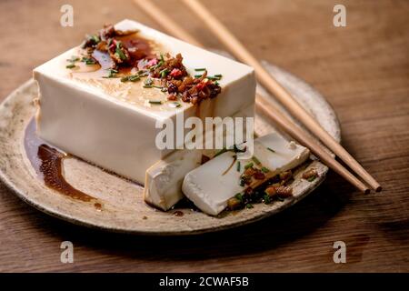 Seidentofu japanischer Sojakäse Vollstück mit Chili, Ingwer und Sojasoße, die auf Keramikplatte mit Essstäbchen über Holztisch bestreichen. Stockfoto