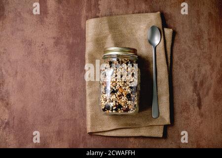 Hausgemachte knackige gebuffte Masergranola aus Hirse mit getrockneten Früchten und Nüssen in Glasmaison-Glas, Löffel auf textiler Serviette über braunem Texturhintergrund. Fl Stockfoto