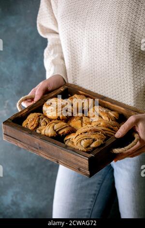 Frau in beigefarbenem Strickpullover in Händen Holztablett mit hausgemahlenen traditionellen schwedischen Kardamom- oder Zimt-Süßbrötchen Kanelbulle Stockfoto