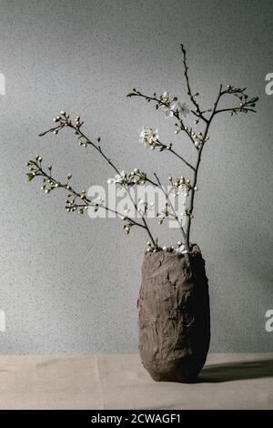 Kirschblüten verzweigt sich in handwerklicher Lehmvase auf grauem Tischtuch. Innendekorationen mit Federdesign. Stockfoto