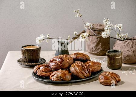 Traditionelle schwedische Zimt süßen Brötchen Kanelbulle auf Vintage-Tablett, Tasse Kaffee, Kanne Sirup, Blütenzweige auf Leinen Tischtuch. Stockfoto