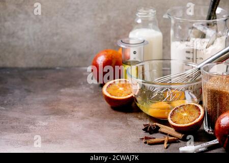 Zutaten für das Backen zu Hause. Mehl, Eier, Rohrzucker, Milch in verschiedenen Schüsseln, Blutorangen und Gewürze. Hintergrund mit grauer Textur Stockfoto