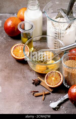 Zutaten für das Backen zu Hause. Mehl, Eier, Rohrzucker, Milch in verschiedenen Schüsseln, Blutorangen und Gewürze. Hintergrund mit grauer Textur Stockfoto