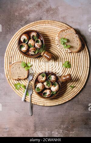 Escargots de Bourgogne - Schnecken mit Kräuterbutter, Gourmet-Gericht, in zwei traditionellen Keramikpfannen mit Petersilie und Brot auf Strohserviette über braunem Tex Stockfoto