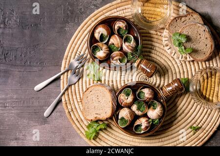 Escargots de Bourgogne - Schnecken mit Kräuterbutter, Gourmet-Gericht, in traditionellen Keramikpfannen mit Petersilie, Brot und ein Glas Weißwein auf Strohnapk Stockfoto