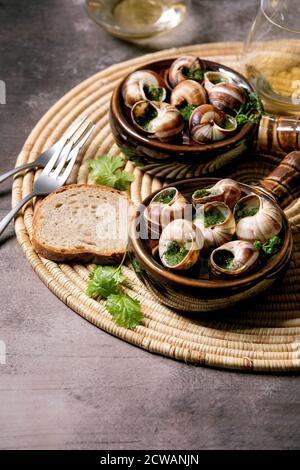 Escargots de Bourgogne - Schnecken mit Kräuterbutter, Gourmet-Gericht, in zwei traditionellen Keramikpfannen mit Koriander, Brot und Glas Weißwein auf stra Stockfoto