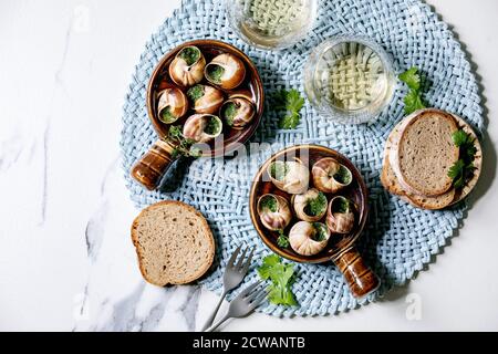 Escargots de Bourgogne - Schnecken mit Kräuterbutter, Gourmet-Gericht, in traditioneller Keramikpfanne mit Koriander, Brot, Glas Weißwein auf blauer Serviette Stockfoto