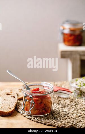 Sonnengetrocknete Kirschtomaten Olivenöl in Glasgefäß mit Gabel, stehend auf Holzküchentisch mit geschnittenem Roggenbrot und grünem Sprosssalat. Stockfoto
