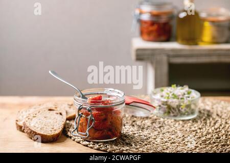 Sonnengetrocknete Kirschtomaten Olivenöl in Glasgefäß mit Gabel, stehend auf Holzküchentisch mit geschnittenem Roggenbrot und grünem Sprosssalat. Stockfoto