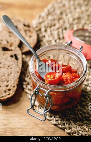 Sonnengetrocknete Kirschtomaten Olivenöl in Glasgefäß mit Gabel, stehend auf Holzküchentisch mit geschnittenem Roggenbrot. Stockfoto