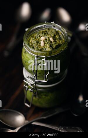 Pesto Genovese - traditionelle italienische grüne Basilikumsoße mit Pinienkernen, Basilikum und Knoblauch in einem Glas mit Löffeln auf rustikalem Holzhintergrund. Medite Stockfoto