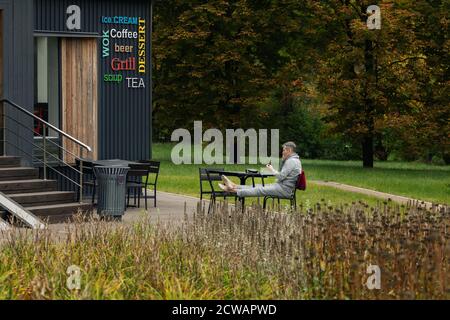 Moskau/Russland - Sep 2020: Älterer kaukasischer Mann sitzt am Kaffeetisch in einem Stadtpark und telefoniert per Kopfhörer. Wolkiger Herbsttag. Stockfoto