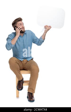 Verwirrt junger Mann am Telefon reden und halten Sprechblase, sitzen isoliert auf weißem Hintergrund, ganzer Körper Stockfoto