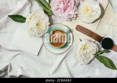 Tasse Espresso Kaffee, leeres Papier, Umschlag, Smart Watch, rosa und weißen Pfingstrosen Blumen mit Blättern über weißem Baumwolltextil Hintergrund. Flach, Stockfoto