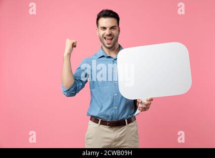 Enthusiastischer Kerl hält Sprechblase und feiert, hält Faust in der Luft und schreit auf rosa Hintergrund Stockfoto