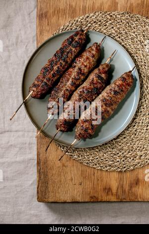 Eastern Fast Food. Gegrilltes, würziges Rindfleisch-Ljulya-Kebab auf Stöcken auf Keramikplatte auf Holztisch mit runder Strohserviette. Flach liegend, Kopierplatz Stockfoto