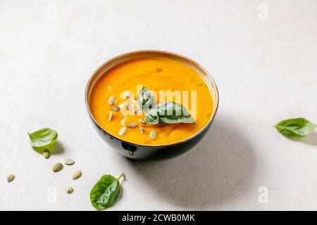 Schüssel mit vegetarischer Kürbis- oder Karotten-Sahnesuppe, dekoriert mit frischem Basilikum, Olivenöl und Kürbiskernen auf weißer Textur-Tabelle mit Zutaten oben. Stockfoto