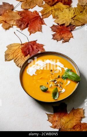 Schüssel mit vegetarischer Kürbis- oder Karotten-Sahnesuppe, dekoriert mit frischen Basilikum-, Sahne- und Kürbiskernen auf weißem Hintergrund mit gelbem Herbstahorn Stockfoto
