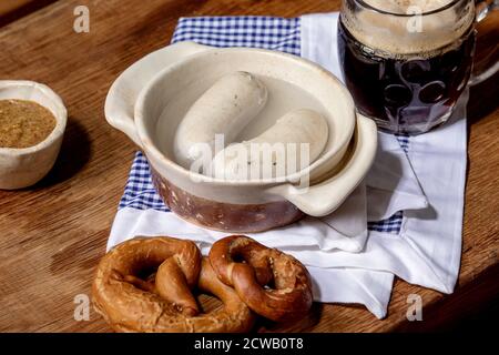 Münchner bayerische traditionelle Weißwürste in Keramikpfanne serviert mit deutschem süßen Senf, einem Becher dunklem Bier und Brezeln Brot auf weißer und blauer Serviette Stockfoto