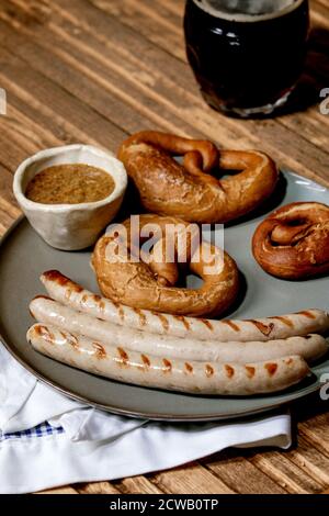 Bayerische traditionelle gegrillte Schweinswürste auf Keramikplatte serviert mit deutschen süßen Senf, Tasse dunkles Bier und Brezeln Brot auf weiß und blau na Stockfoto