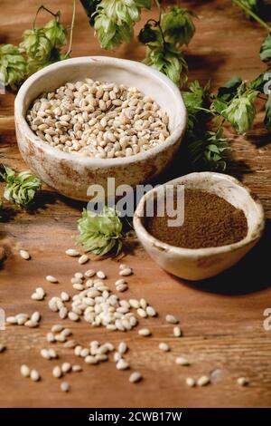 Alles zum Brühen. Frische grüne Hopfenzapfen, Weizenkorn und rotfermentiertes Malz in Keramikschalen auf Holzhintergrund. Stockfoto