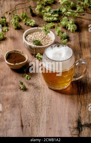 Klassischer Glas-Becher mit frischem, kalt schaumigem Lagerbier mit grünen Hopfenzapfen, Weizenkorn und rotfermentiertem Malz in Keramikschalen dahinter über Holzstruktur Stockfoto