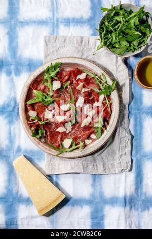 Klassischer Rindercarpaccio mit Käse und Rucola auf Keramikplatte, serviert mit Olivenöl über weißer und blauer Tischdecke. Flach liegend, Platz Stockfoto