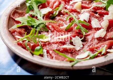 Klassischer Rindercarpaccio mit Käse und Rucola auf Keramikplatte über weißer und blauer Tischdecke. Stockfoto