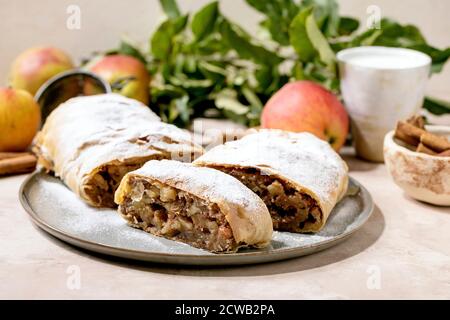 Hausgemachter klassischer Apfelstrudel in Scheiben mit Puderzucker auf Keramikplatte mit frischen Äpfeln, grünen Blättern und Zimtstangen darüber. Stockfoto