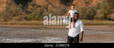 Kleiner Junge, der auf dem Nacken ihres Vaters sitzt. Schöner junger afroamerikanischer Vater und sein süßer Sohn. Banner, Panorama, Kopierbereich. Vater und Sohn verbringen Zeit zusammen Stockfoto