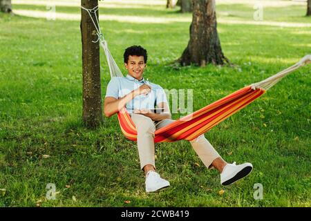 Lächelnder Kerl, der in der Hängematte im Park sitzt und digital hält Tablet Stockfoto