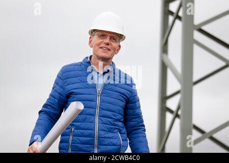 Lächelnder Ingenieur mit Hut und bluepriint in der Hand Stehen vor einem elektrischen Pol - selektiver Fokus Auf die Person Stockfoto