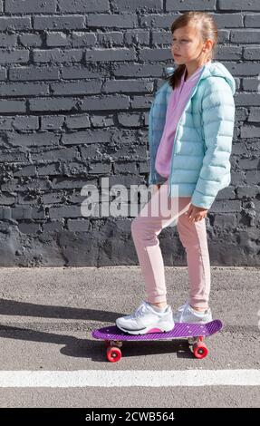 Kleines Mädchen in bunten Kleidern mit einem Skateboard steht in der Nähe grauen Ziegelwand, vertikale Foto Stockfoto