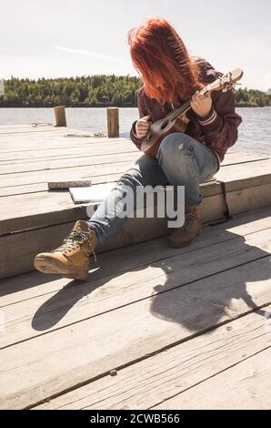 Rothaarige Teenager-Mädchen singt ein Lied mit Ukulele sitzen Auf einem hölzernen Pier an sonnigen Tagen Stockfoto