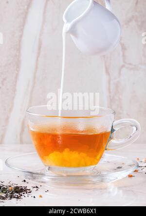 Milch gießen aus weißem Krug Glas Tasse heißen Tee auf Untertasse mit trockener grüner und schwarzer Tee Blätter über weißen Marmor Backgtound. Stockfoto