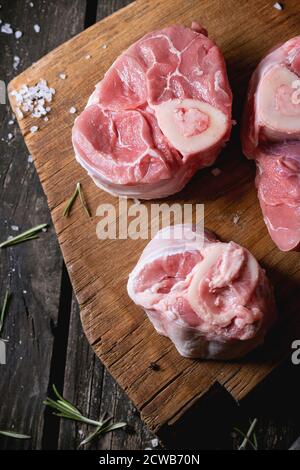 Fleisch, roh Osso Buco auf Holzbrett mit Salz, Pfeffer und Rosmarin über alten Holztisch. Dunkel rustikalen Stil. Stockfoto