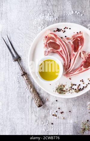 Rohe Lammkoteletts mit Salz, Pfeffer, trockenen Kräutern und einer Schüssel Olivenöl auf weißem Keramikteller über weißem Holztisch. Fleischgabel in der Nähe. Draufsicht. Stockfoto