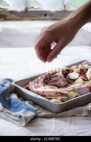 Frau Handsalze marinierte rohe Lammkoteletts mit Zwiebel und Pfeffer in alten Aluminium-Backform. Über Küchentisch mit weißer Tischdecke. Natürlicher Tag Stockfoto