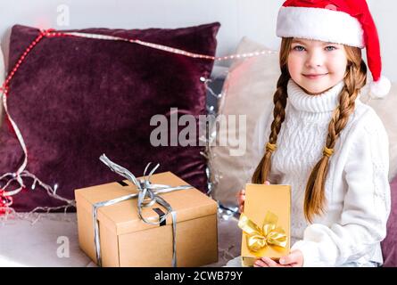 Ein Mädchen in einem weihnachtsmann Hut sitzt auf dem Sofa mit einem Neujahr Geschenk in ihren Händen und Schaut auf die Kamera Stockfoto