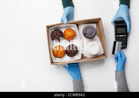 Moderne kontaktlose Bezahlung für Bestellung. Kurier in Gummihandschuhe gibt Terminal und Box mit Muffins und Kaffee, Client wendet Kreditkarte Stockfoto