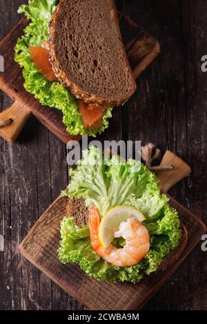 Sandwich mit Vollkornbrot, frischem Salat, Shrimps und gesalzenem Lachs auf kleinem Holzbrett über altem Holztisch. Rustikaler Stil. Draufsicht Stockfoto