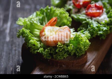 Sandwich mit Vollkornbrot, frischem Salat, Garnelen, Feta-Käse und Kirschtomaten auf Holzschneidebrett über altem Holztisch. Dunkler rustikaler Stil Stockfoto