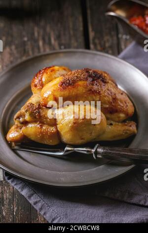 Ganze gegrillte Mini Huhn auf Vintage Metallplatte über alten Holztisch. Mit Gabel, Serviette und Gewürzen. Dunkel rustikalen Stil. Stockfoto