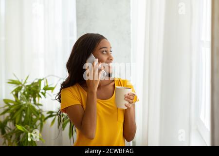 Wunderschöne afroamerikanische Dame in der Nähe Fenster mit Tasse Kaffee und Kommunikation auf dem Smartphone zu Hause Stockfoto