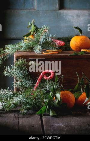 Mandarinen mit Blättern im Weihnachtsdekor mit Weihnachtsbaum, trockener Orange und Bonbons auf Holzkiste über altem Holztisch. Dunkler rustikaler Stil Stockfoto