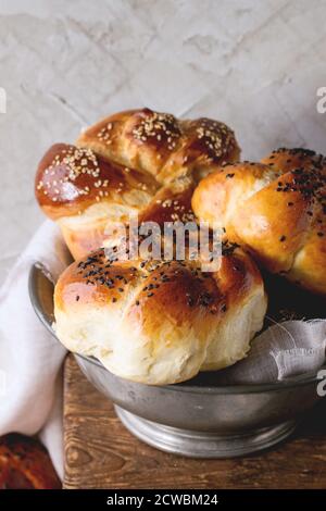 Haufen süße runde Sabbat challah Brot mit weißem und schwarzem Sesam in Vintage metall Schüssel auf Holztisch mit verputzten Wand im Hintergrund. Stockfoto