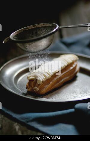 Single Eclair mit weißer Buttercreme in vintage Metallplatte mit Sieb Zucker Pulver. Dunkler rustikaler Stil. Tageslicht. Außenansicht Stockfoto