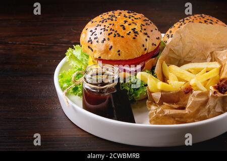 Frisch hausgemachter Hamburger mit schwarzen Sesamkörnern auf weißem Teller mit pommes Frites Kartoffeln, serviert mit Ketchup-Sauce im Glas über dunklem Holz-Ta Stockfoto